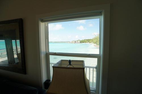 Staniel Cay Vacation Rentals, One bedroom on the water with view of the Staniel Cay Yacht Club. View through a window showing a beach with clear turquoise water, sky with few clouds, and a lamp on the windowsill indoors.