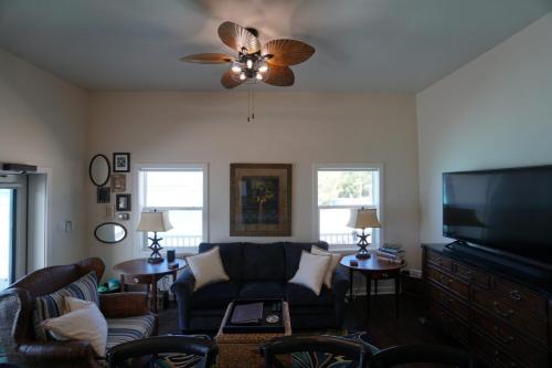 Staniel Cay Vacation Rentals, One bedroom on the water with view of the Staniel Cay Yacht Club. Living room with a dark blue sofa, wicker chair, large TV, ceiling fan, two side tables with lamps, and wall art, facing two large windows showing an outside view.