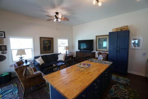 Staniel Cay Vacation Rentals, One bedroom on the water with view of the Staniel Cay Yacht Club. A living room with blue cabinets, a wooden island, sofas, wicker chairs, and a flat-screen TV. Framed art hangs on cream walls, and a ceiling fan with lights is overhead.